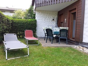 Um pátio com duas poltronas de descanso e uma mesa, cercado por um sebedo e um edifício com parede branca texturizada e porta de madeira.