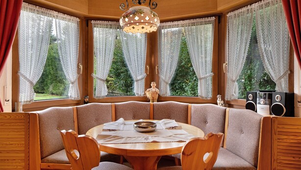 A wooden dining area with a round table, cushioned benches, lace curtains, and a decorative ceiling light. - Apartment 'Nr 2' with Private Terrace, Private Garden and Wi-Fi (Titisee-Neustadt)