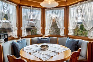 Ein gemütliches Zimmer mit einem runden Tisch, gepolsterten Sitzgelegenheiten, Spitzenvorhängen und Blick durch große Fenster.