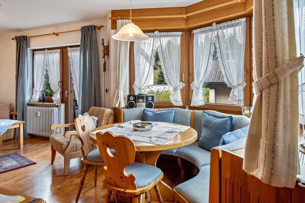 Een gezellige woonkamer met een ronde tafel, houten stoelen, een bank en grote ramen met gordijnen.