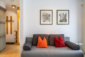 Uma sala de estar moderna com um sofá cinzento, dois almofadas laranja e uma vermelha, uma mesa de centro de vidro e duas obras de arte emolduradas na parede.