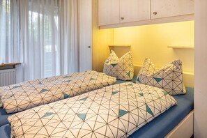 Quarto com cama, almofadas, janela com cortinas, radiador e armário de madeira.