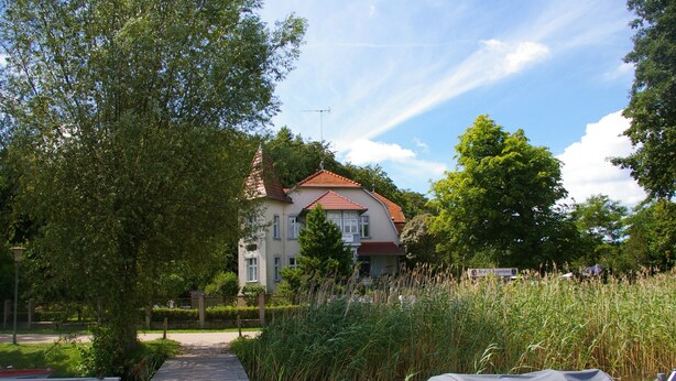 Puinen laituri johtaa punaiseksi maalattuun taloon, jonka ympärillä on vehreää kasvillisuutta ja rauhallinen vesialue.
