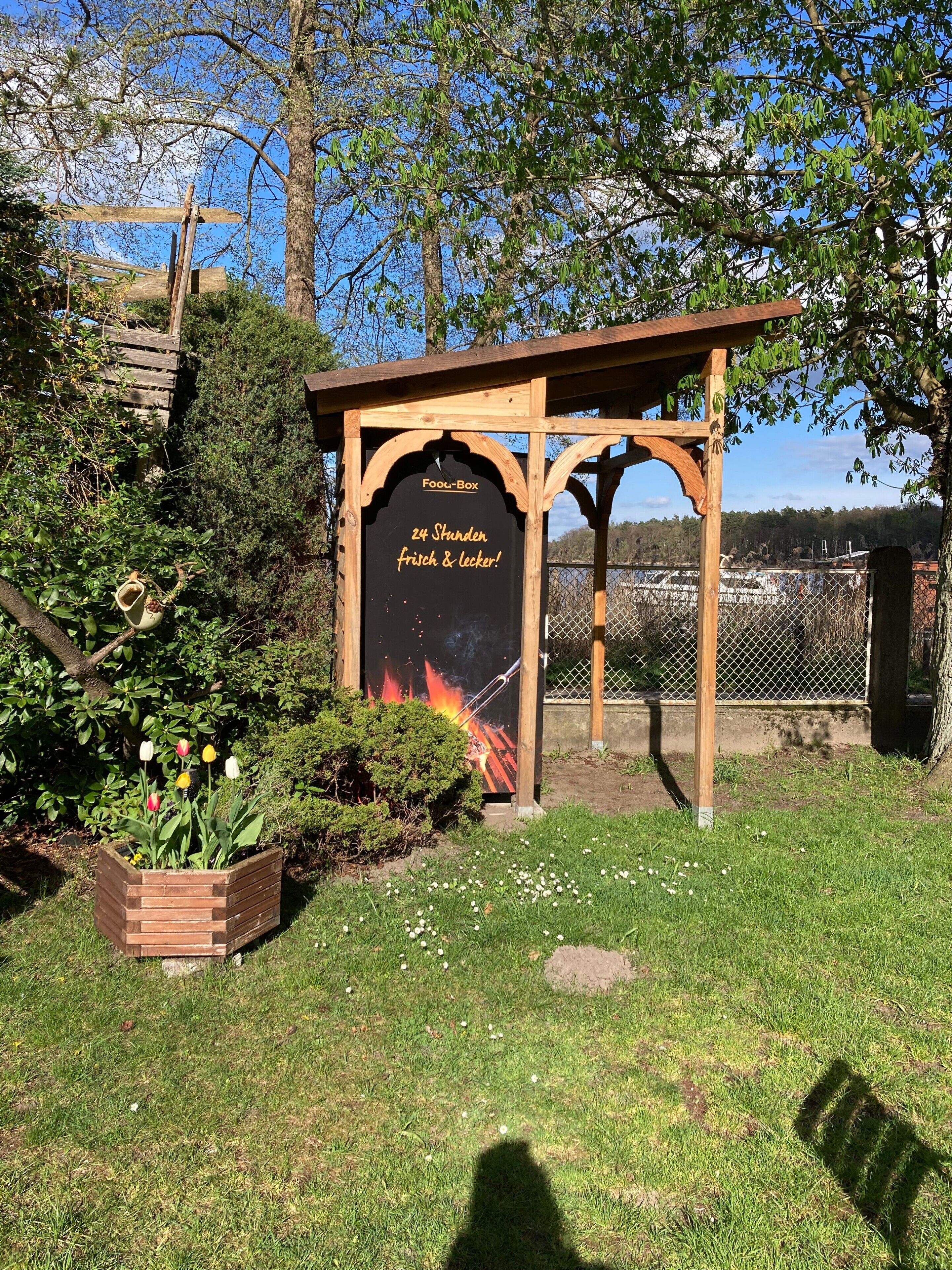 A wooden structure labeled 'Fool-Box' with a sign stating '24 Stunden frisch & lecker!' surrounded by greenery and a fenced area.