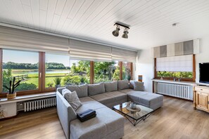Sala de estar moderna com sofá seccional, mesa de centro de vidro e grandes janelas com vista para uma paisagem verde.