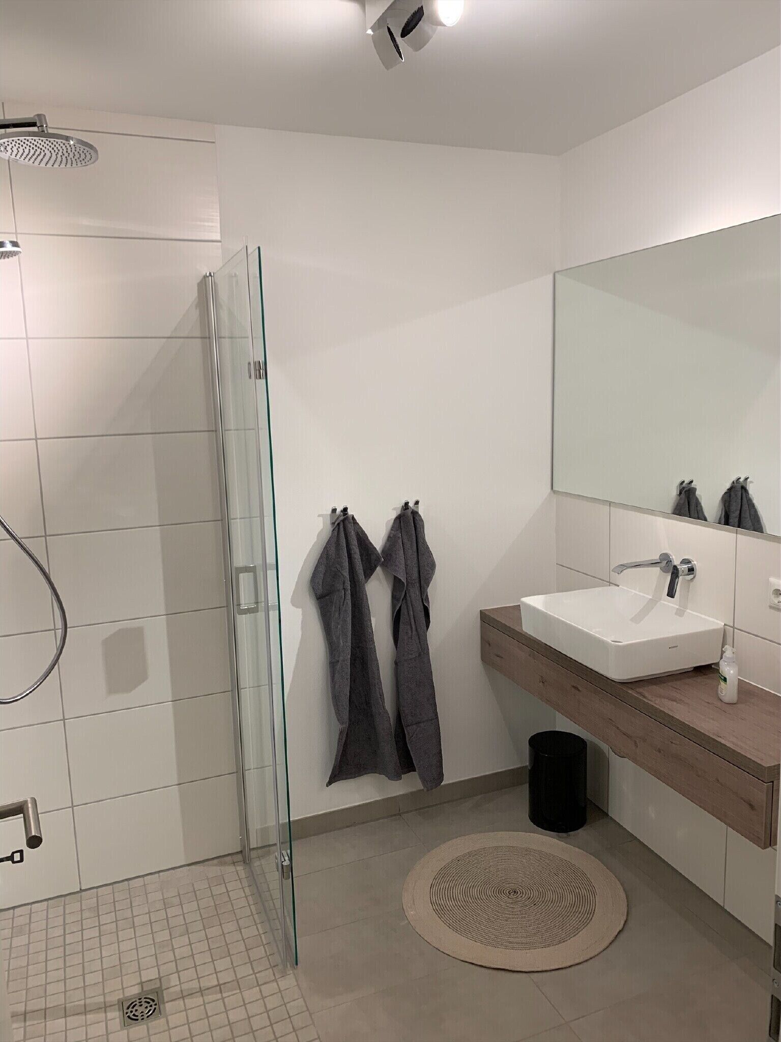 Uma casa de banho moderna com cabine de duche em vidro, lavatório com espelho e um tapete redondo no chão.