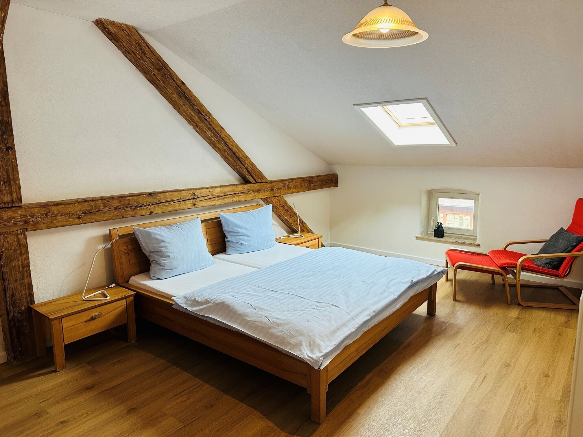 A bedroom with a wooden bed, a nightstand, a red chair, a skylight, and a window.