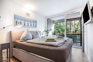 Une chambre à coucher avec un lit, une table de chevet, une lampe, un tableau et une fenêtre donnant sur des arbres.