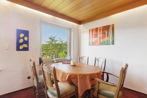 Un coin repas comprenant une table ronde, six chaises, une fenêtre donnant sur un paysage verdoyant et deux tableaux accrochés aux murs.