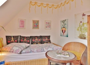 Une pièce chaleureuse avec un lit, un fauteuil en osier, une petite table et des œuvres d’art accrochées au mur.
