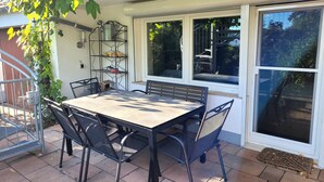 Une terrasse avec une table et des chaises, une étagère en métal avec divers objets, et une vue à travers une fenêtre.