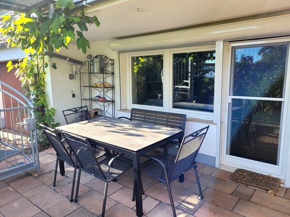 Une terrasse avec une table et des chaises, une étagère en métal avec divers objets, et une vue à travers une fenêtre.