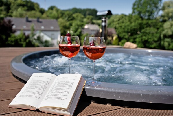 An open book on a wooden deck, two glasses of red drink with cherries, and a hot tub in the background.