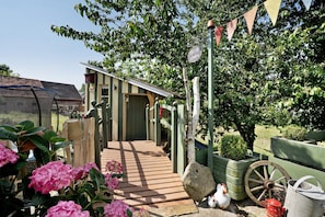 Un patio in legno conduce a una casa con una porta verde, immersa nel verde e circondata da un giardino.