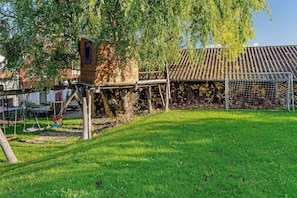 Une maisonnette en bois, des balançoires et un but de football dans une cour verdoyante, avec un toit en tuiles en arrière-plan.