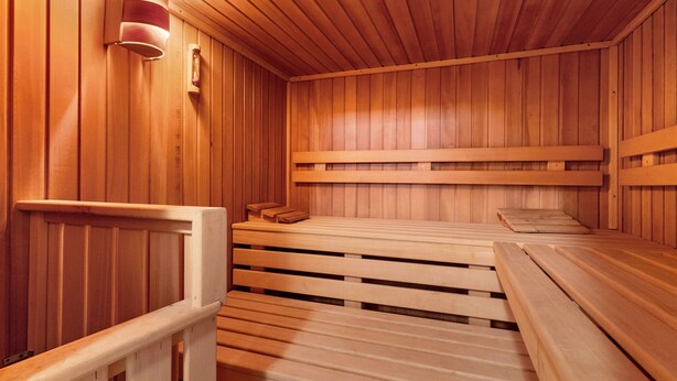 A wooden sauna with benches and a bench seat, wooden walls, and a wall-mounted light fixture.