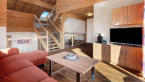 Un intérieur en bois avec un escalier, un canapé, une table sur laquelle se trouve un gâteau, et un téléviseur posé sur un meuble en bois.
