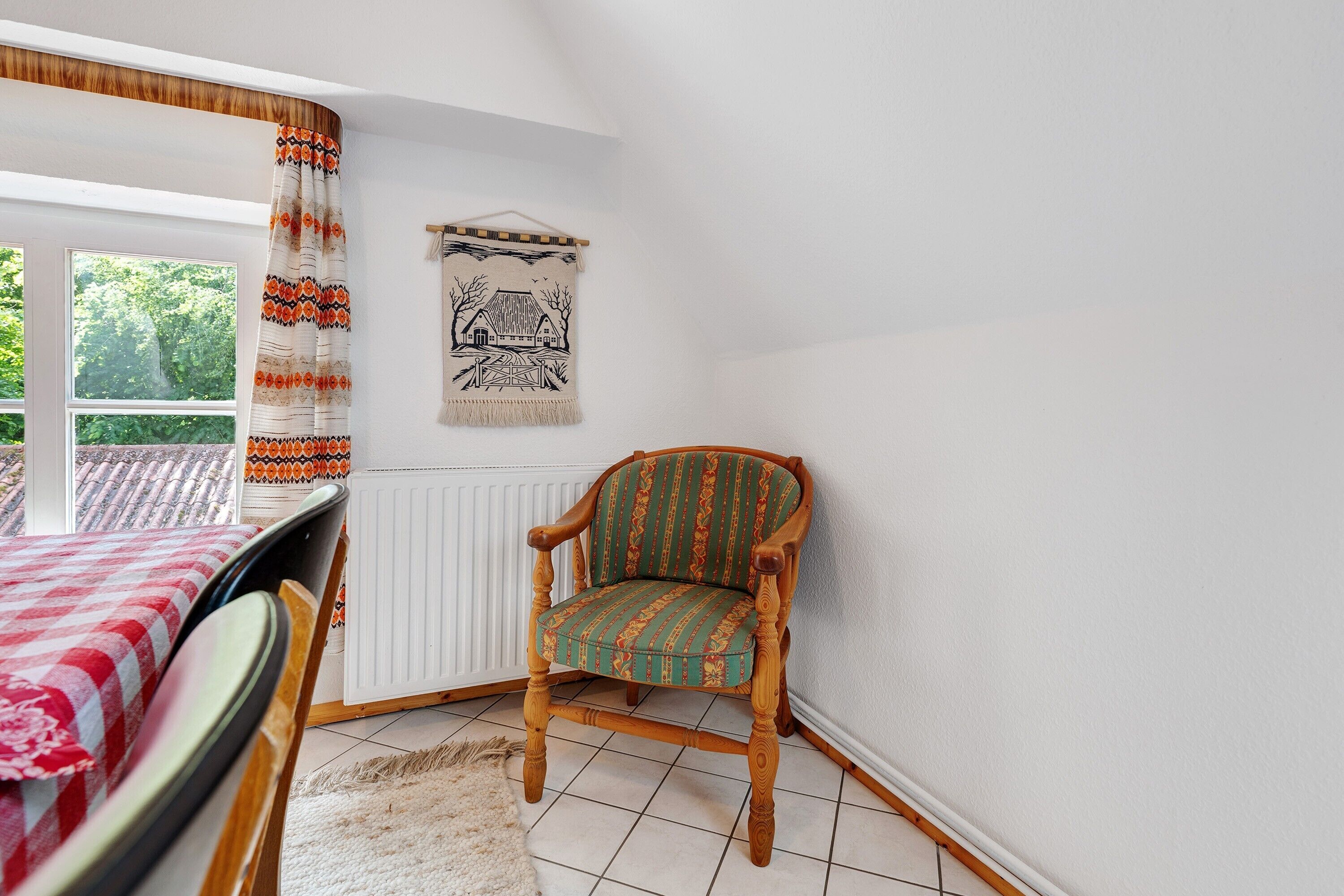 A room with a wooden chair, a table with a red and white checkered tablecloth, a white radiator, and a wall tapestry.
