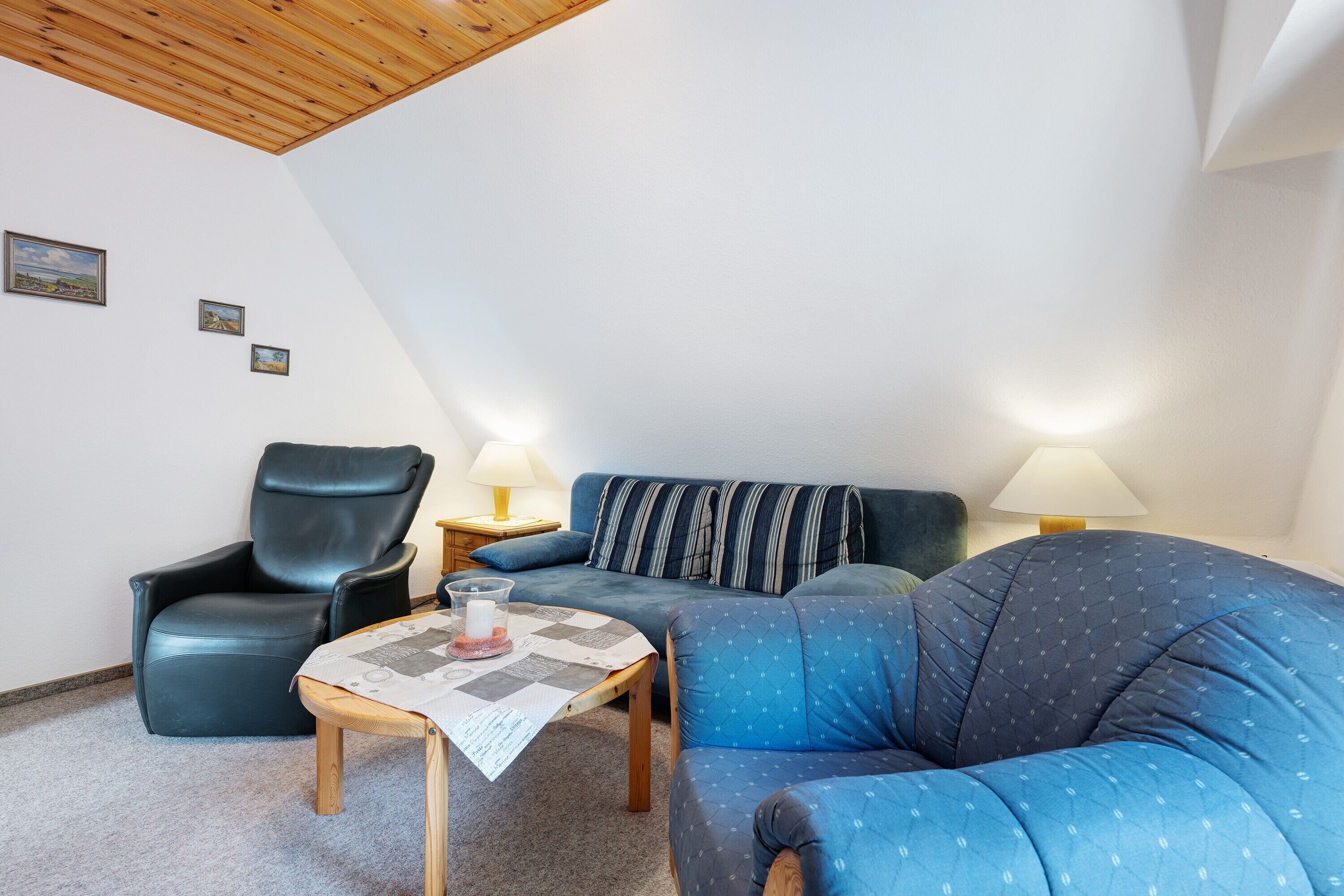 A living room with a wooden ceiling, a blue sofa, a black leather chair, a small table with a placemat, and two bedside lamps.