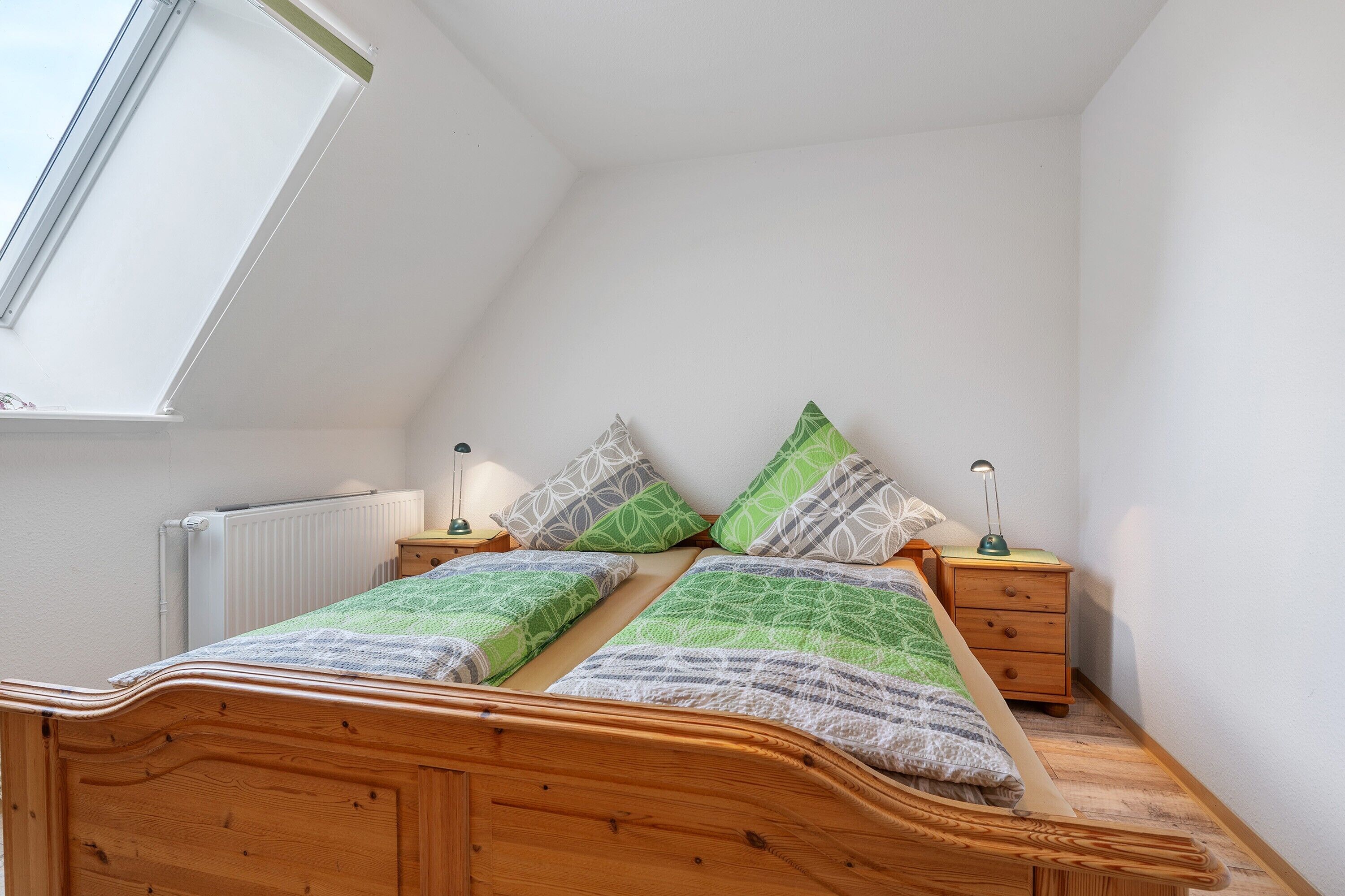 A bedroom with a sloped ceiling, a wooden bed, two bedside tables with lamps, and a radiator.