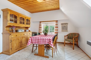 A dining area with a wooden cabinet, a table with a checkered tablecloth, chairs, a potted plant, and a window with curtains.