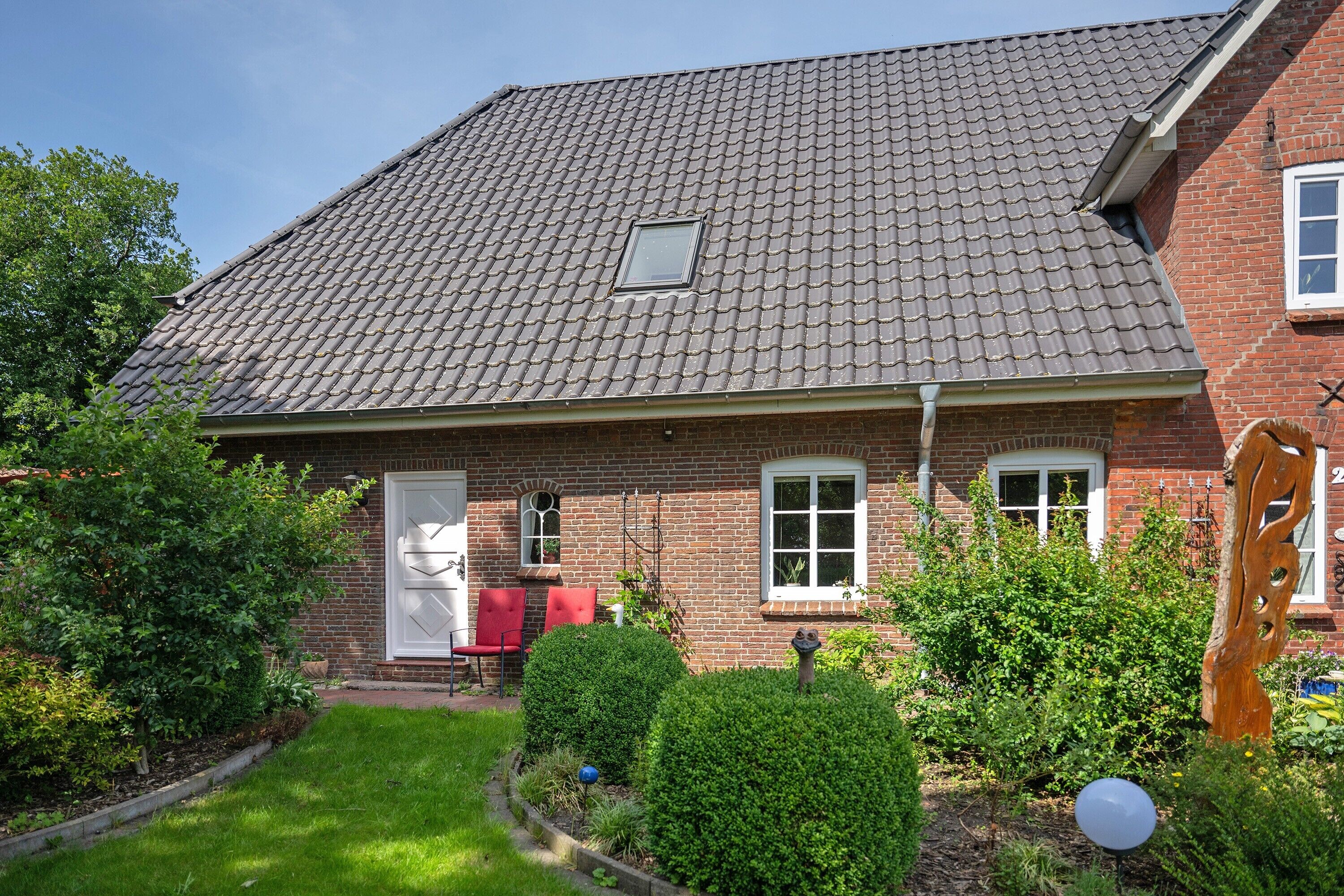 A brick house with a tiled roof, a small window, and a garden with hedges and a wooden sculpture.