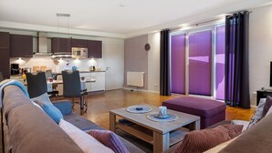A modern living room with a sofa, a coffee table, and a dining area with chairs and a kitchen in the background. - Apartment 'Fewo Birkenallee - Lavendel' with Private Terrace (Deinste)