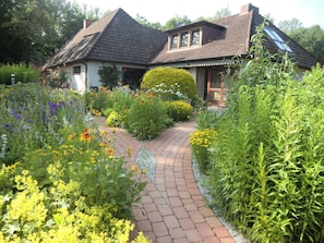Een huis met een pad van bakstenen dat naar de ingang leidt, omringd door diverse bloeiende planten en struiken.