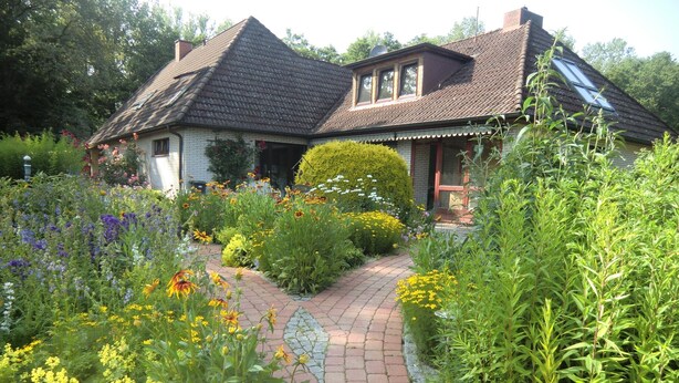 Een huis met een pad van bakstenen dat naar de ingang leidt, omringd door diverse bloeiende planten en struiken.
