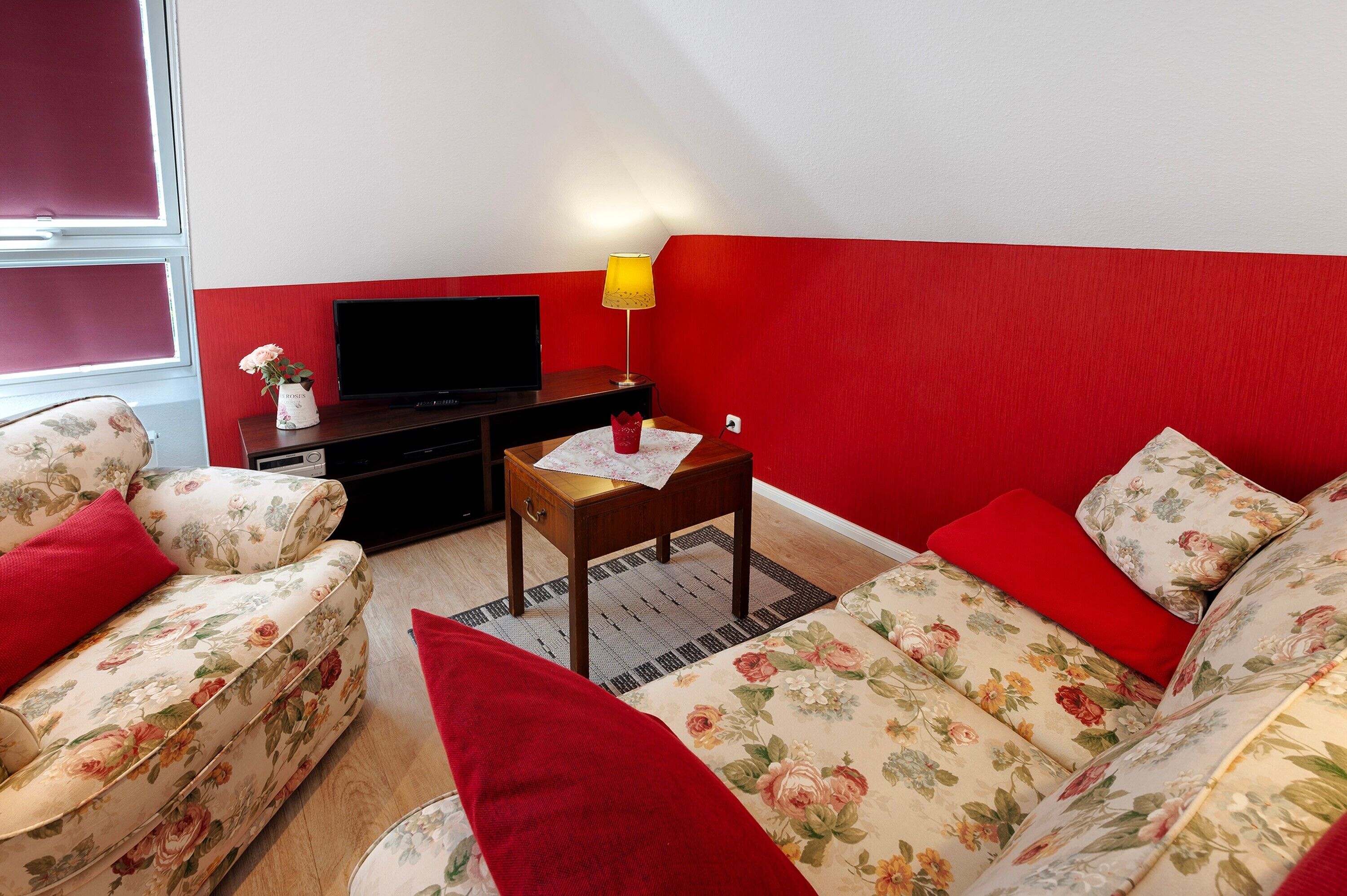 Un salon avec des canapés à motifs floraux, un téléviseur à écran plat, une petite table en bois avec une lampe et un mur d’accent rouge.