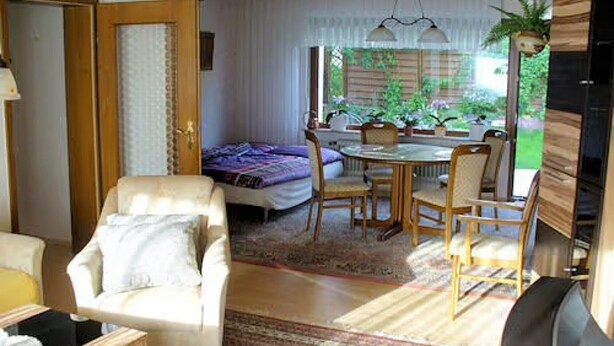 Een kamer met een bed, een stoel, een tafel en een eethoek met stoelen. Er is een raam met gordijnen en uitzicht op groen buiten.