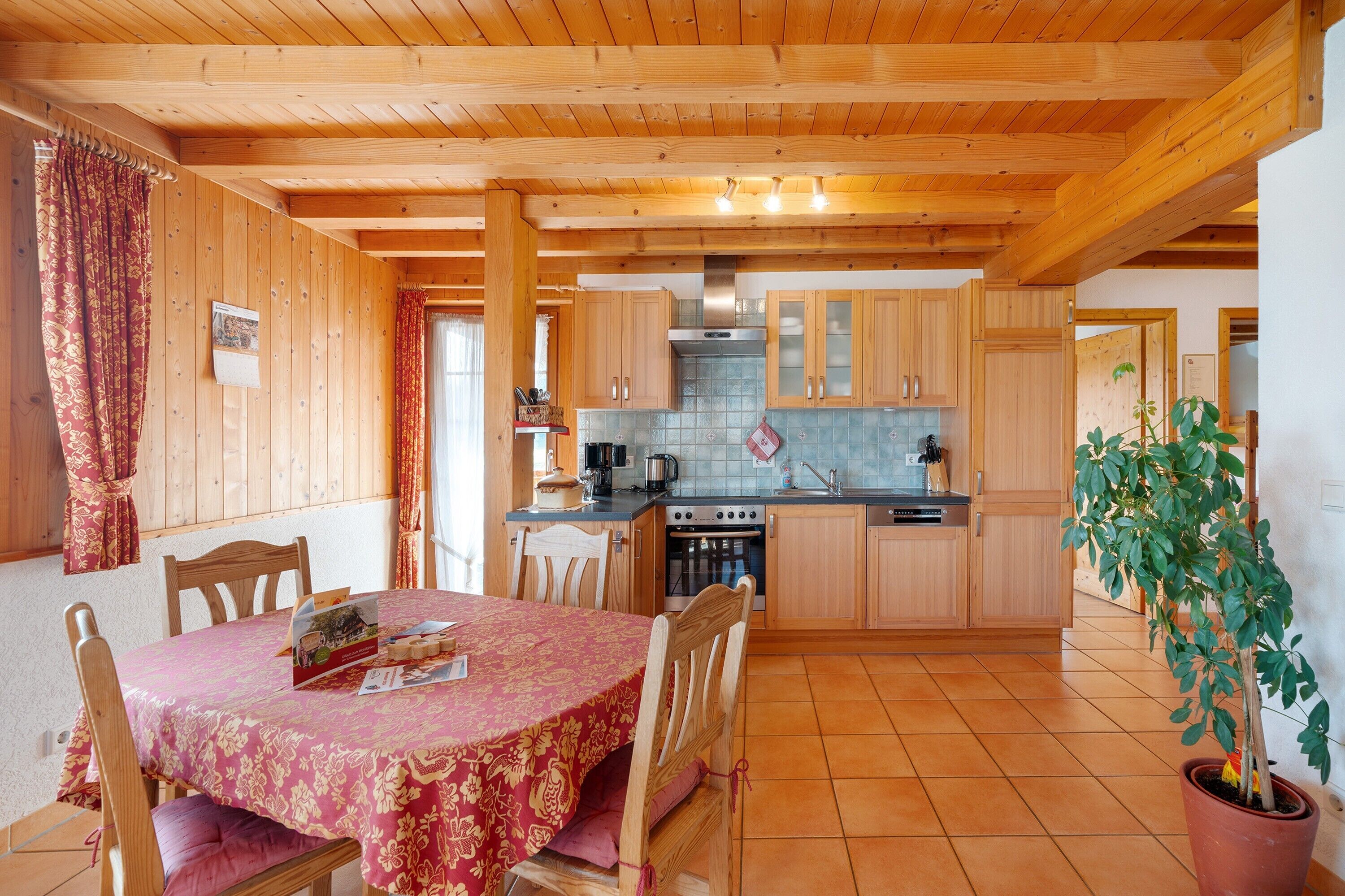 Un interior de madera con una mesa de comedor, sillas, una zona de cocina con armarios y electrodomésticos, y una planta en maceta.