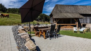 A patio with a wooden table and chairs, a large umbrella, and a rustic wooden building in the background. - Mühlen Chalet Apartment with Private Terrace (Titisee-Neustadt)