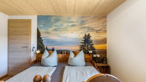 A bedroom with a wooden headboard, two beds, a nightstand, a wall-mounted lamp, and a mural of a sunset with trees. - Mühlen Chalet Apartment with Private Terrace (Titisee-Neustadt)