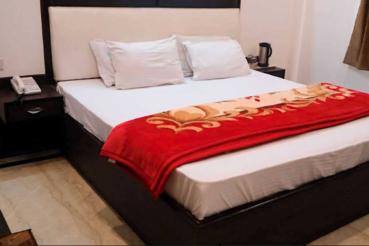A hotel room with a bed, white pillows, a red blanket, a bedside table with a phone, and a nightstand.