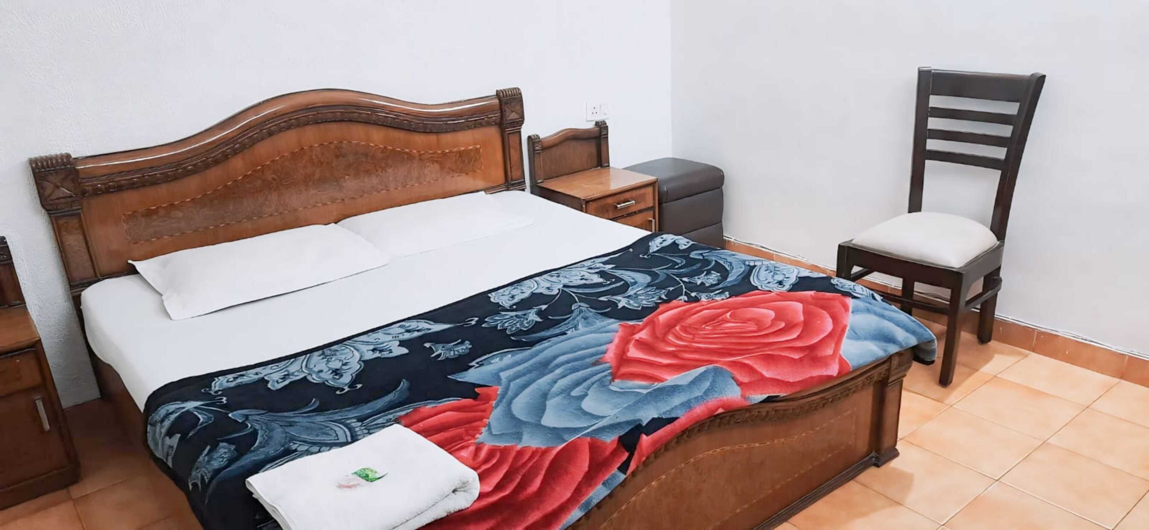 A neatly made bed with a floral bedspread, a wooden nightstand with a lamp, a chair, and a small table with a lamp.