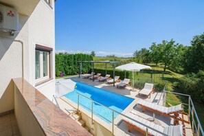 A rooftop pool with lounge chairs and umbrellas, surrounded by greenery and a clear sky.
