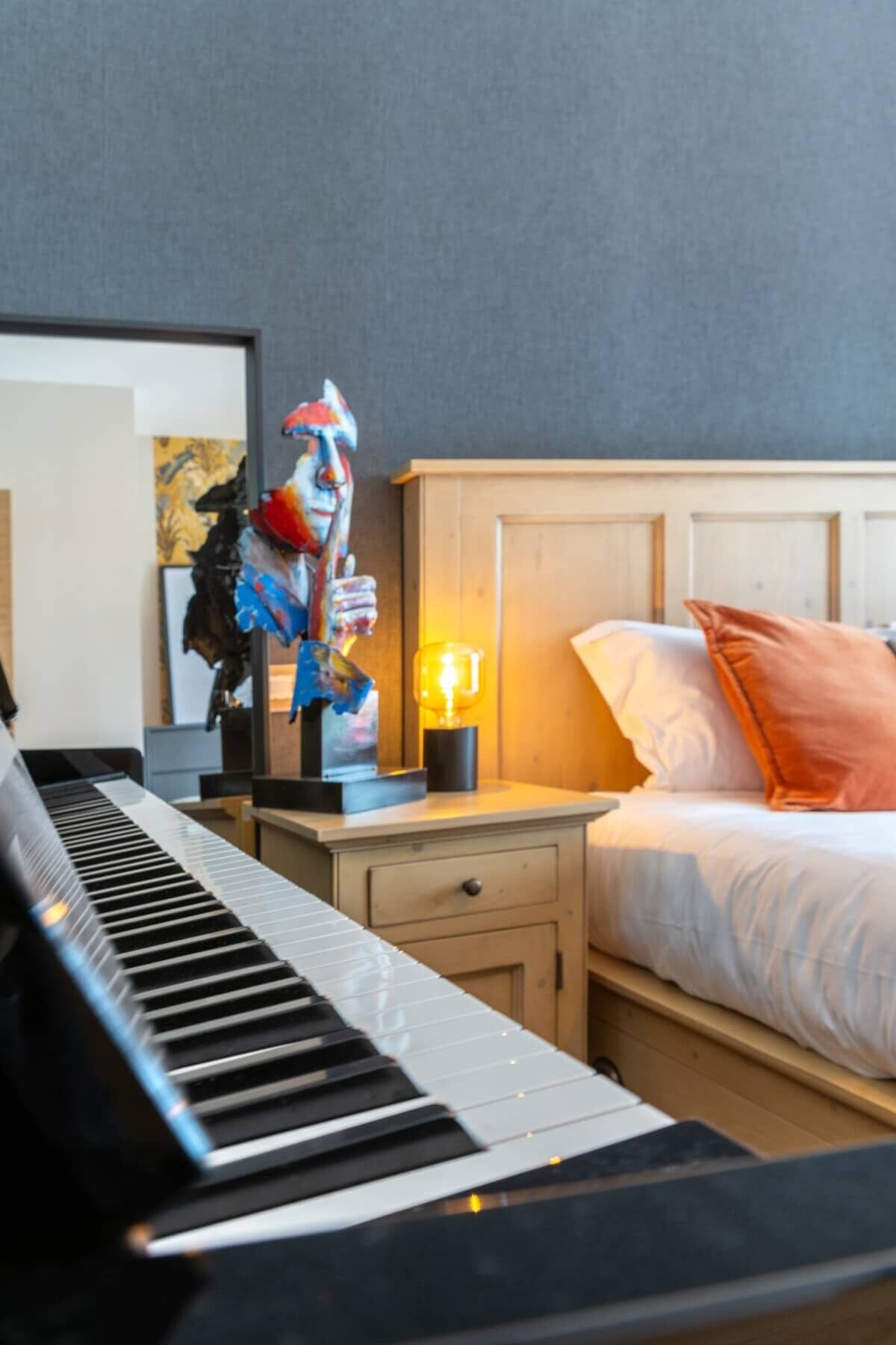Habitación de hotel con un piano, una cama con ropa de cama blanca y naranja, una mesita de noche con una lámpara y un cuadro colgado en la pared.