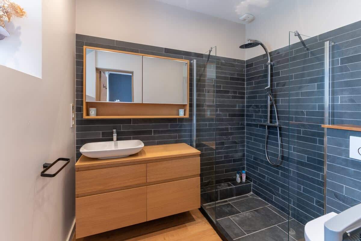 A modern bathroom with a wooden vanity, a large mirror, and a shower area with dark tiles.
