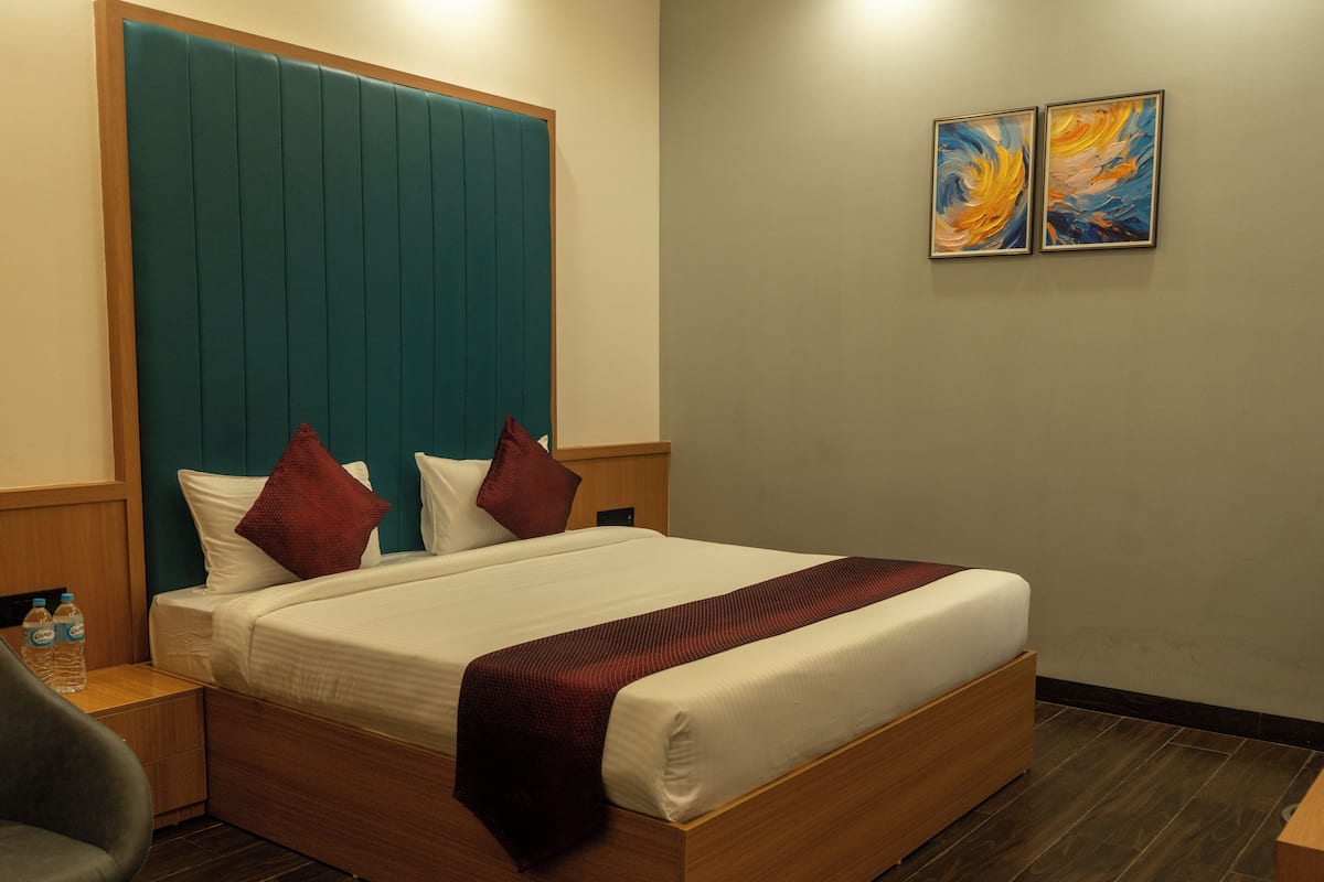 A hotel room with a large bed, wooden headboard, two pillows, a bedside table with a bottle, and two paintings on the wall.