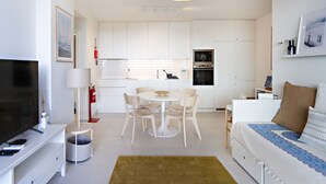 Una sala de estar moderna y bien iluminada con un televisor de pantalla plana, una mesa de comedor redonda con sillas, un sofá con un cojín estampado, y una zona de cocina con armarios y electrodomésticos blancos.