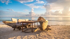 Una playa con una mesa y sillas de madera, un cubo grande de madera y un mar tranquilo al fondo.