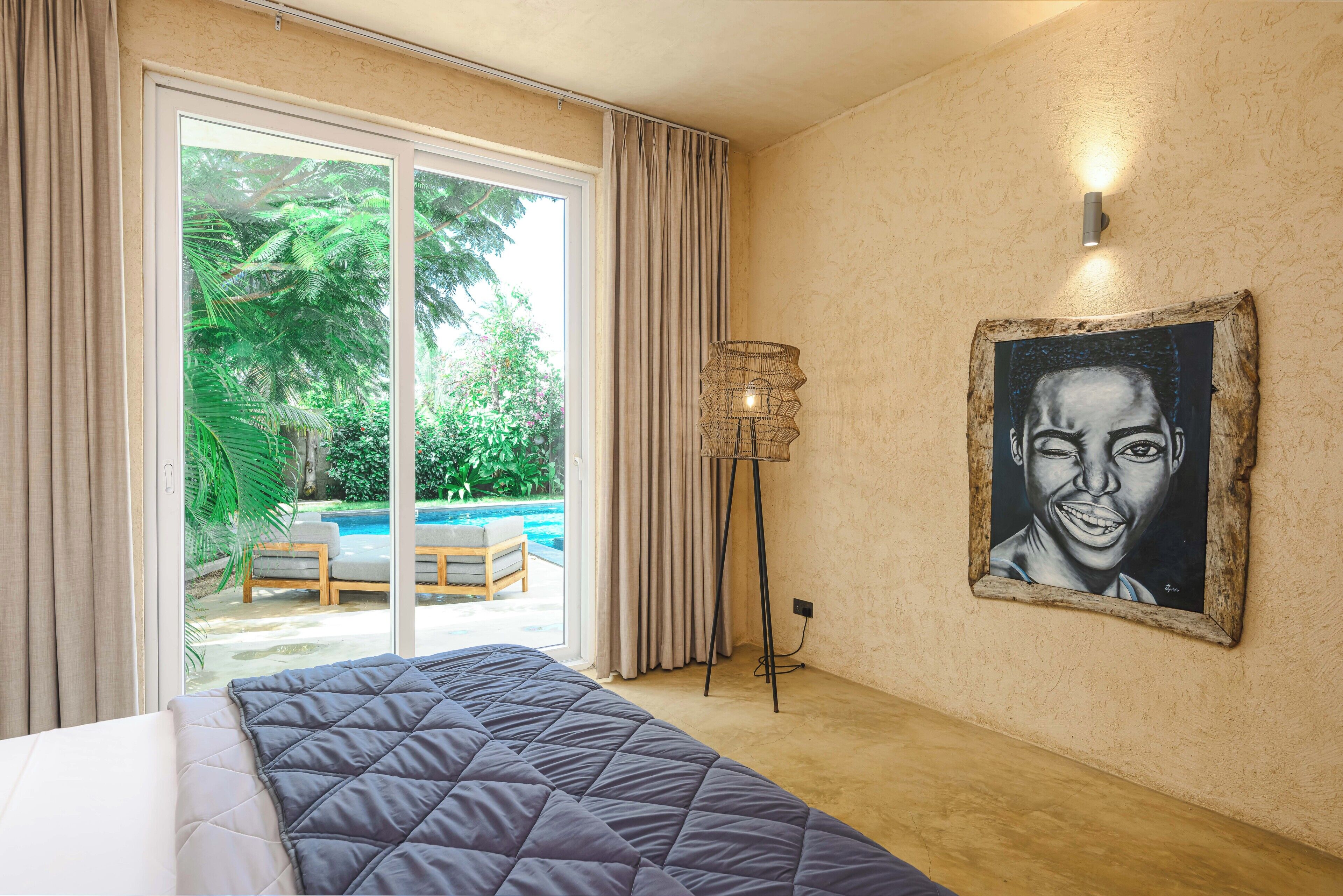 Habitación de hotel con cama, un cuadro, lámpara de pie y vista a una piscina exterior y vegetación.