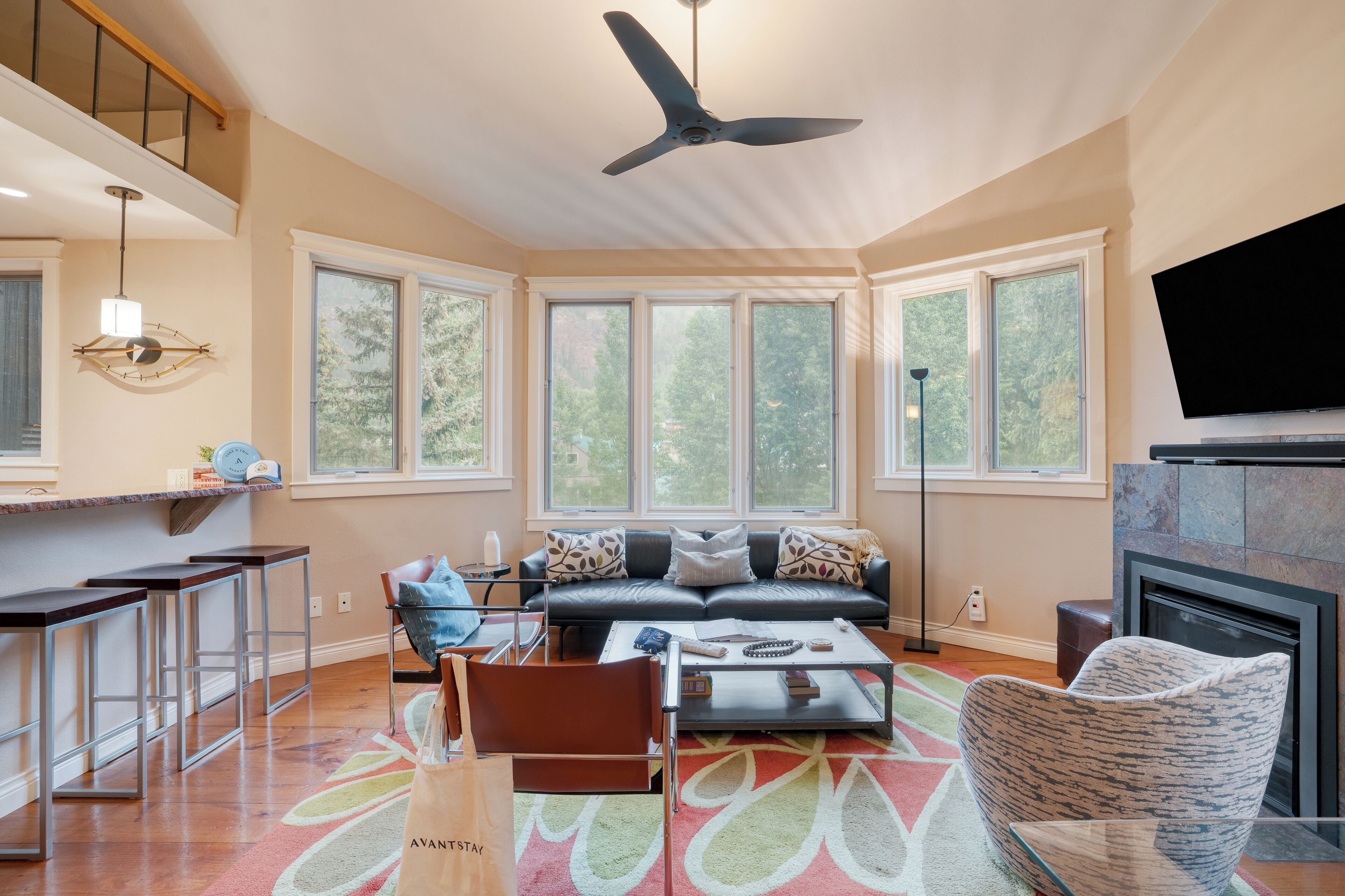 A spacious living room with a large window, a ceiling fan, a fireplace, and a bar area.
