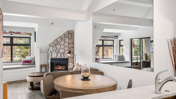 A modern living room with a stone fireplace, a round dining table, and a kitchen area with a sink and window. - Lake Placid Lodge Loft 2 bedroom (Whistler)