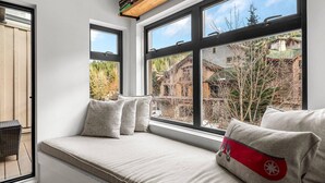 A cozy seating area with a cushioned bench and pillows, large windows offering a view of a wooded area and buildings. - Lake Placid Lodge Loft 2 bedroom (Whistler)