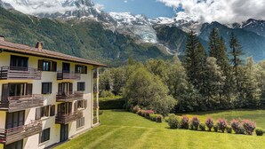 A two-story building with balconies, surrounded by a well-maintained lawn and flowering bushes, with a mountain range in the background.