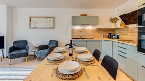 A modern dining area with a wooden table set for four, a kitchen with a built-in oven, and a seating area with two armchairs.