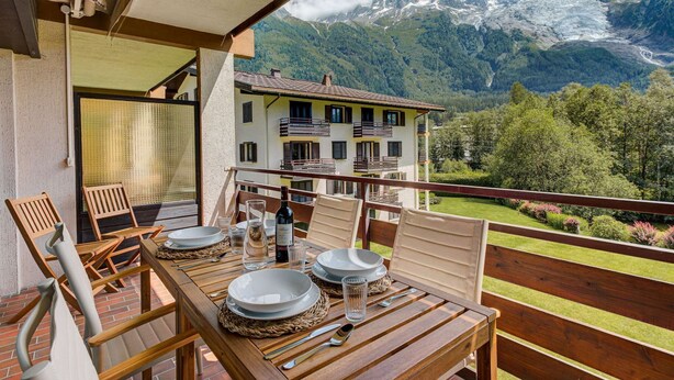 A balcony with a wooden table set for dining, overlooking a mountain landscape with a chalet in the background.