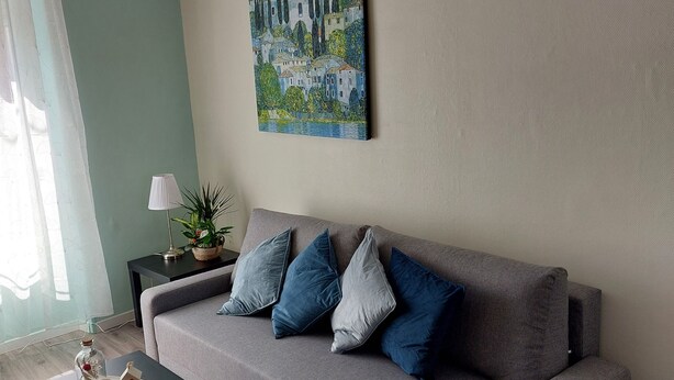 A living room with a grey sofa, a black coffee table, a painting on the wall, and a ceiling-mounted air conditioner. - Véro's Lodges. Duplex 5 minutes from Verdun, private parking, garage for two-wheeled vehicles, air conditioning (Thierville-sur-Meuse)
